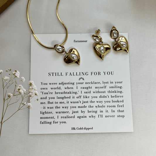A gold-tone necklace and earrings set with a heart charm, accompanied by a note and displayed on a white surface with some dried flowers.