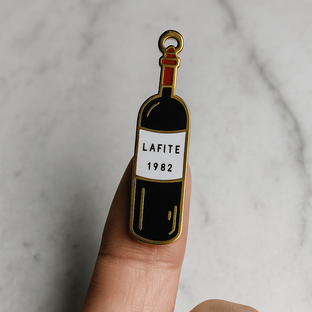 Enamel pin of a wine bottle labeled 'Lafite 1982' held between fingers on a marble background