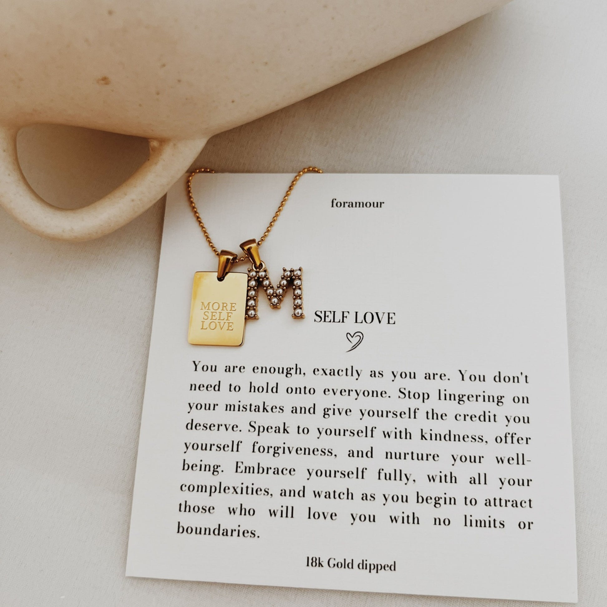 Gold necklace with 'M' pendant on a card with text and a mug in the background