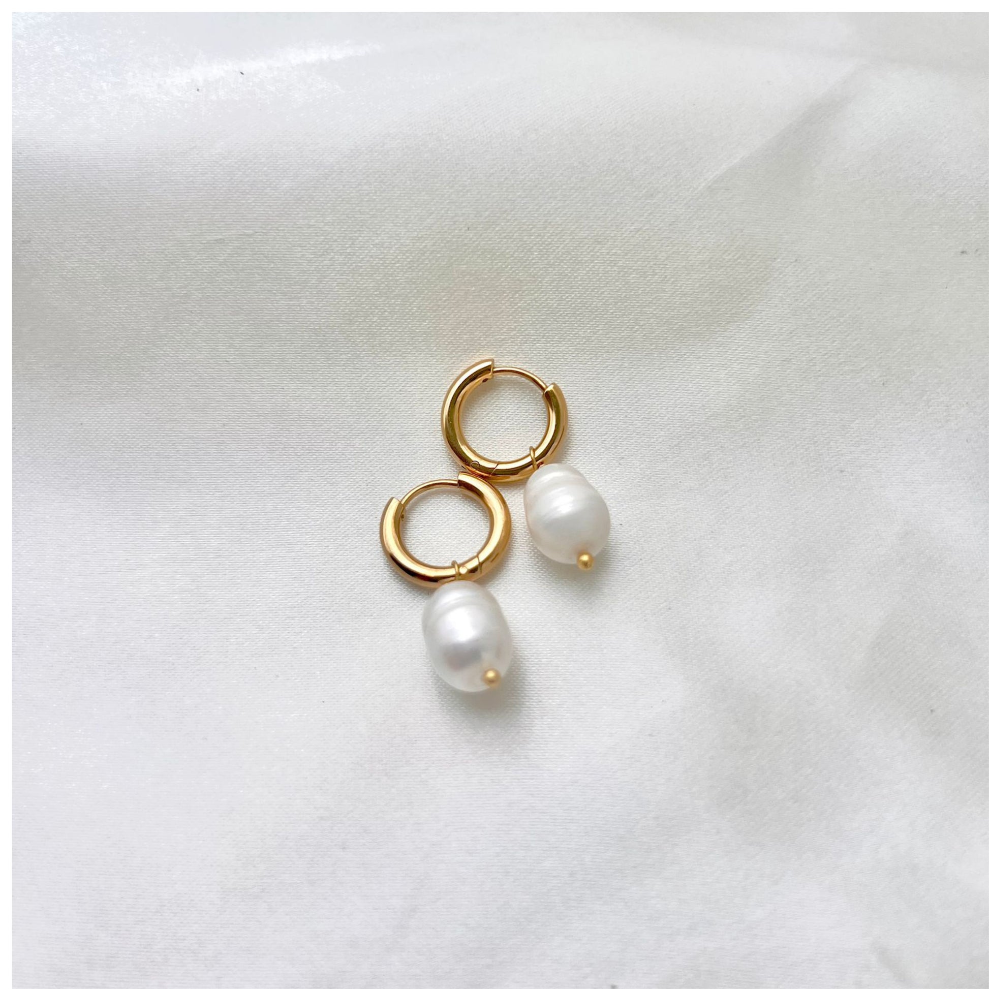 A pair of gold hoop earrings with white pearls attached, placed on a white fabric surface.
