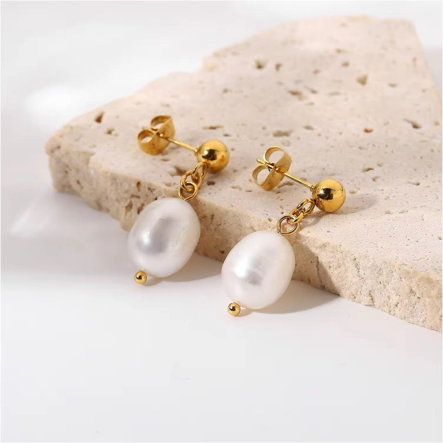 A set of 18kt gold plated earrings featuring a fresh water pearl drop, displayed on a beige stone surface.