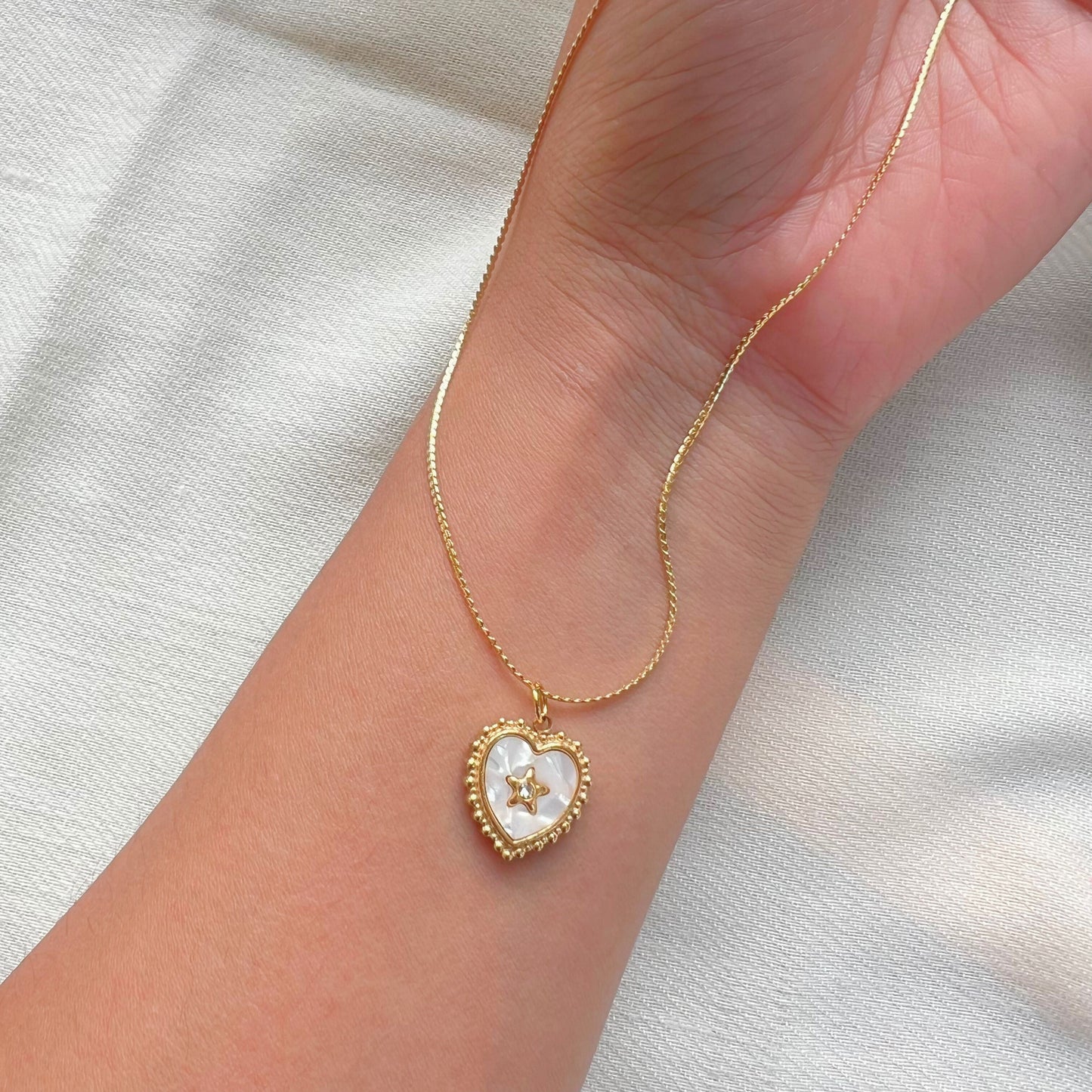 A gold-colored necklace with a heart-shaped pearl pendant in the center, worn on a person's wrist.