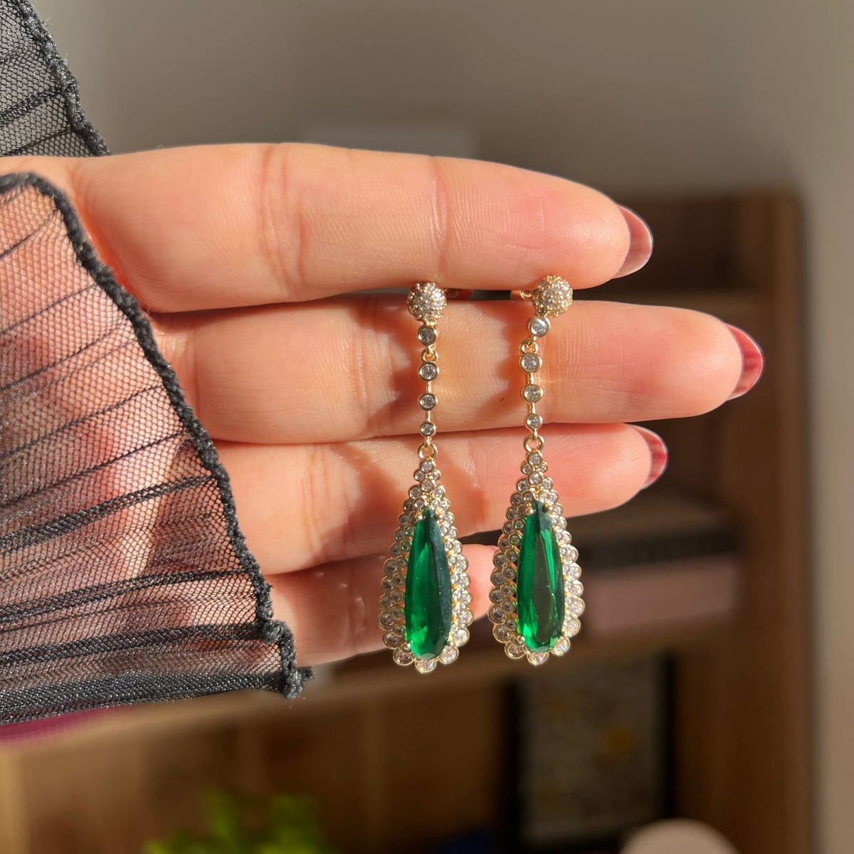 Green teardrop earrings held between fingers with a blurred background