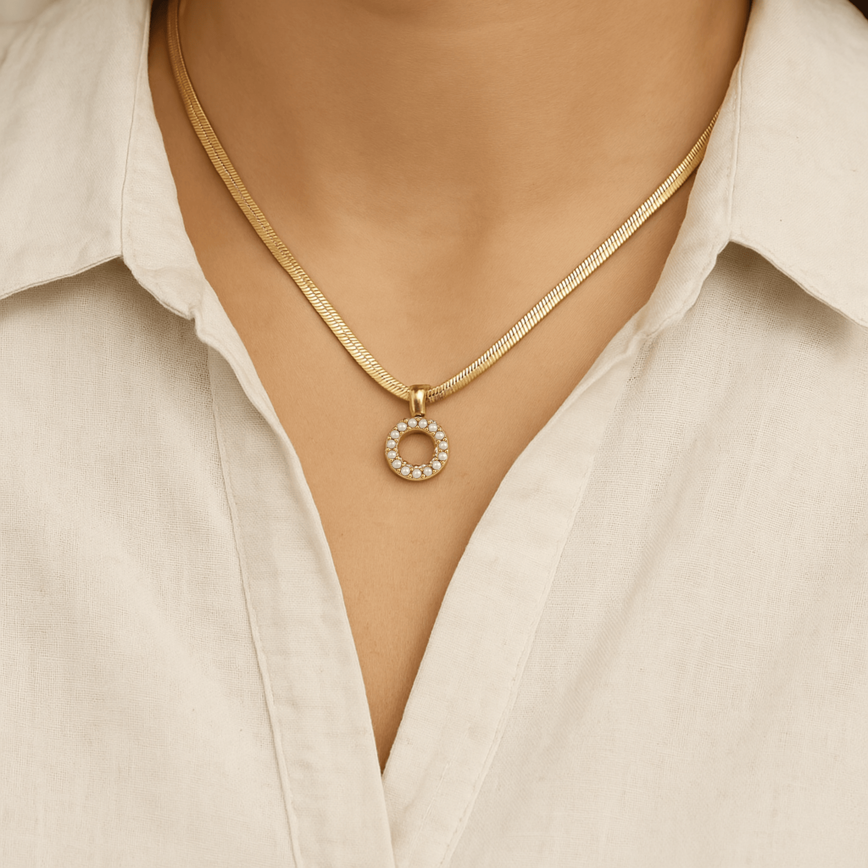A gold-colored necklace with a circular charm pendant worn by a person with a white shirt.
