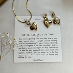 A gold-tone necklace and earrings set with a heart charm, accompanied by a note and displayed on a white surface with some dried flowers.