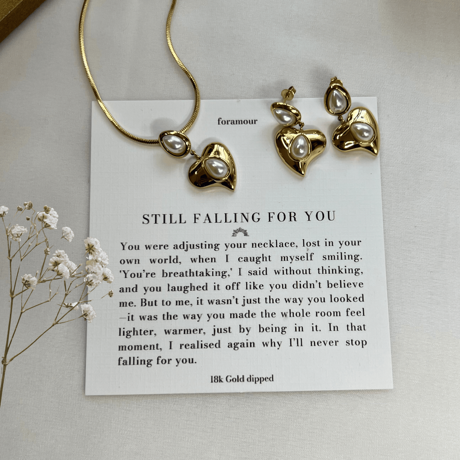 A gold-tone necklace and earrings set with a heart charm, accompanied by a note and displayed on a white surface with some dried flowers.