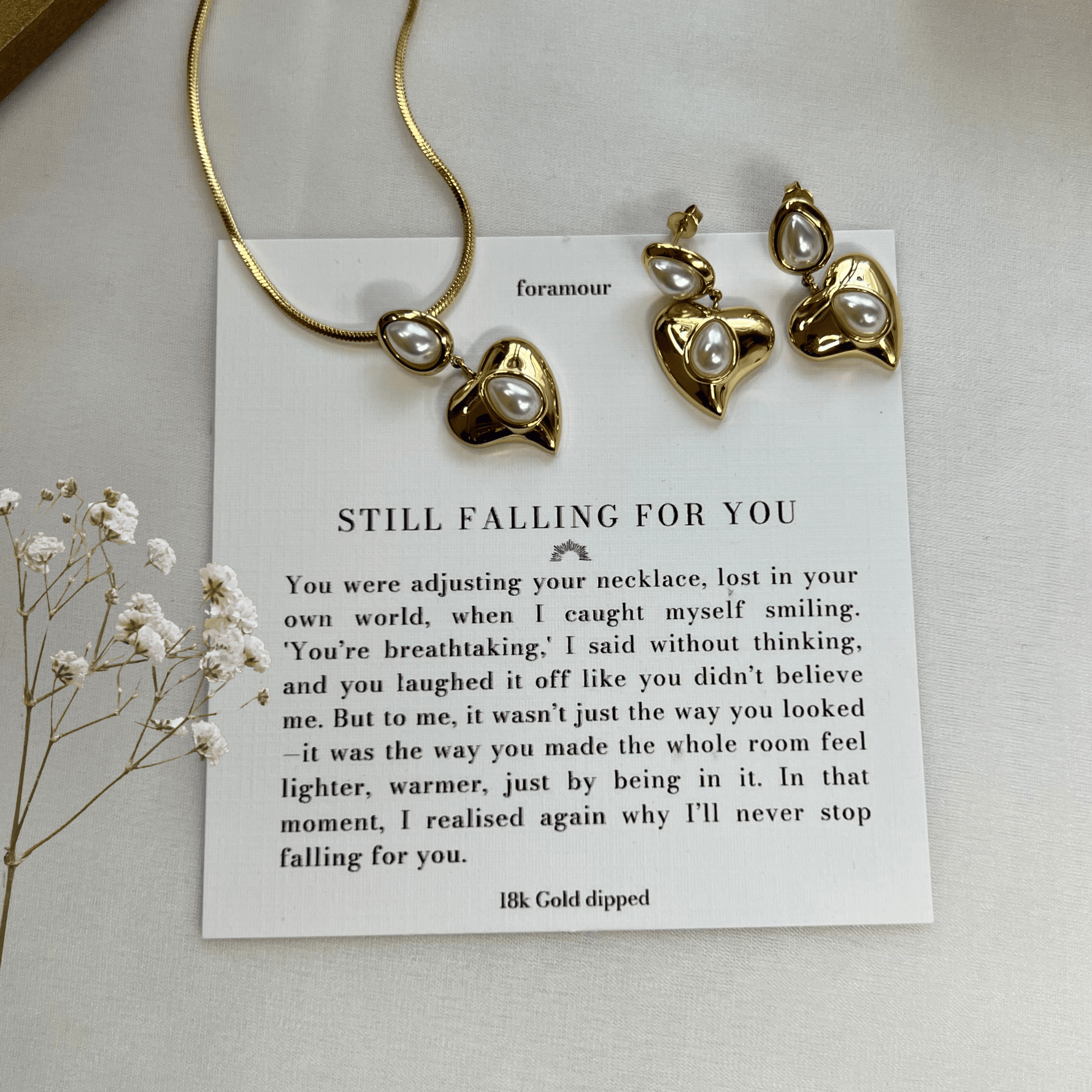 A gold-tone necklace and earrings set with a heart charm, accompanied by a note and displayed on a white surface with some dried flowers.