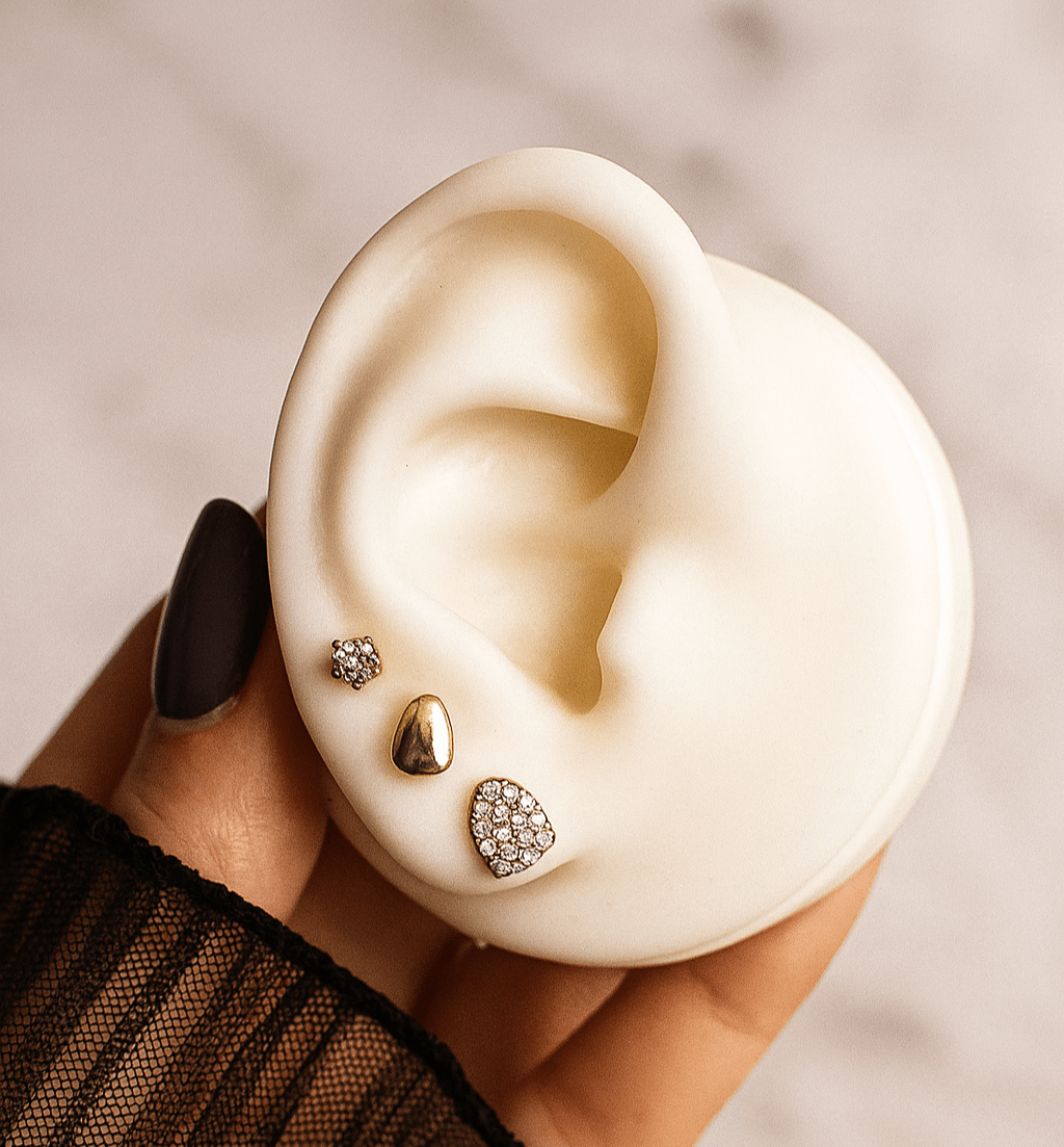 Close-up of an ear with three earrings held by a hand against a neutral background