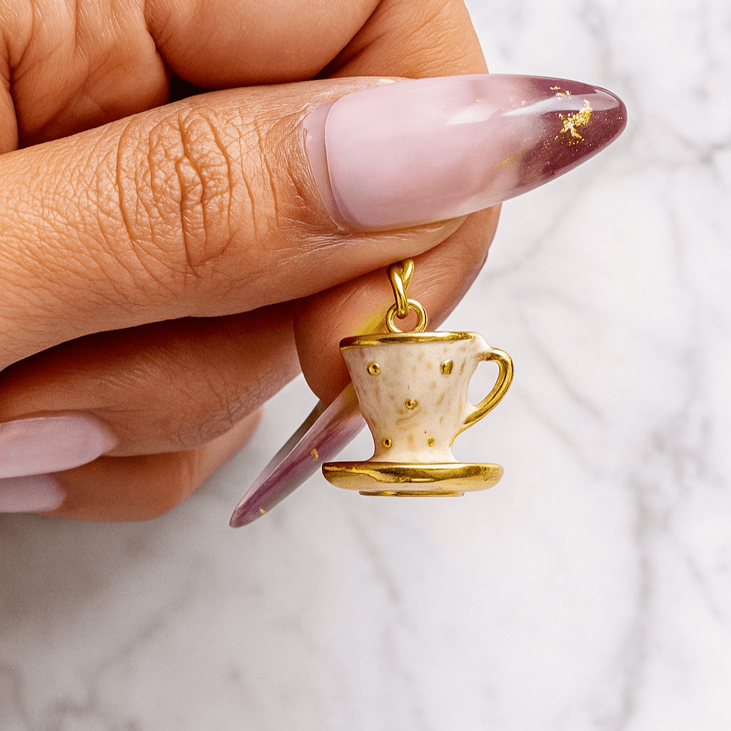 Gold teacup charm held by a hand with pink nail polish on a marble background