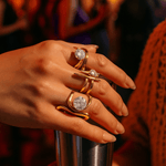 Hand wearing gold rings with gemstones against a blurred background of people and lights