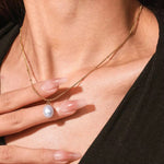 A gold-colored necklace with a delicate beaded chain and a single white pearl pendant, displayed against a white background with a small floral design to the side.