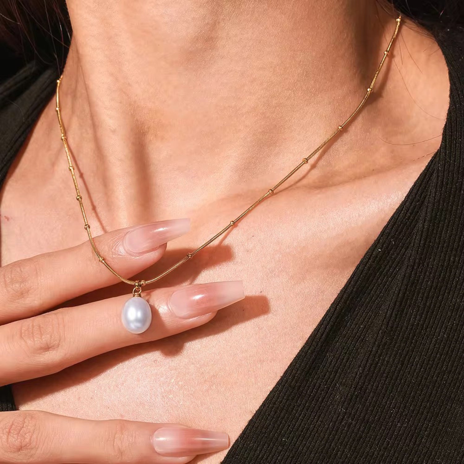 A gold-colored necklace with a delicate beaded chain and a single white pearl pendant, displayed against a white background with a small floral design to the side.