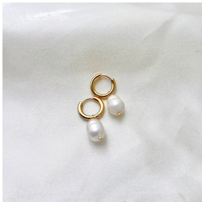 A pair of gold hoop earrings with white pearls attached, placed on a white fabric surface.
