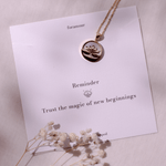 An 18k gold-dipped necklace with a floral design pendant, displayed on a card that reads 'Reminder Trust the magic of new beginnings' along with some dried flowers and the brand name 'Foramour'.