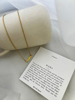A gold-tone necklace with the word 'Mamía' written on a card, presented next to a cylindrical object, possibly a gift box.