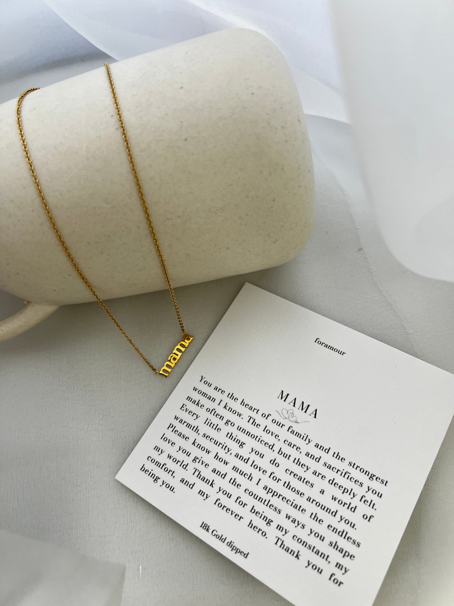 A gold-tone necklace with the word 'Mamía' written on a card, presented next to a cylindrical object, possibly a gift box.
