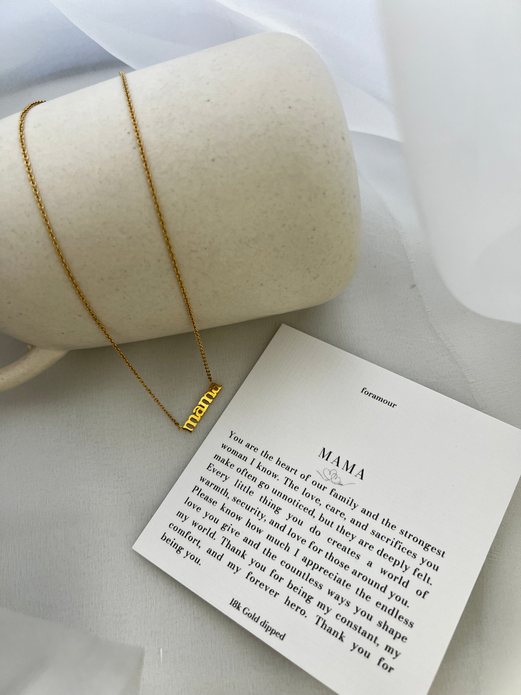 A gold-tone necklace with the word 'Mamía' written on a card, presented next to a cylindrical object, possibly a gift box.