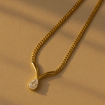 A gold-colored necklace with a single teardrop-shaped crystal pendant, displayed against a beige background.