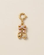 Gold charm shaped like ballet shoes with a pink bow on a beige background