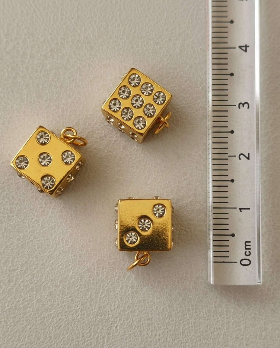 Gold dice-shaped charms with embedded stones on a gray surface with a ruler for scale.