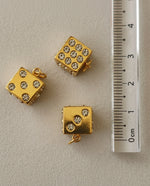 Gold dice-shaped charms with embedded stones on a gray surface with a ruler for scale.