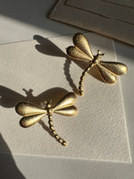 Gold dragonfly-shaped earrings on a textured beige surface