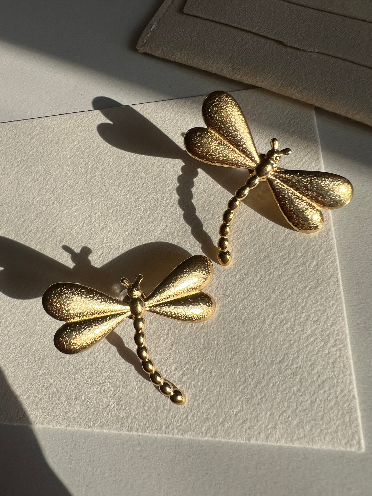 Gold dragonfly-shaped earrings on a textured beige surface