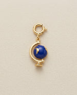 Gold globe charm with a blue gemstone on a beige background