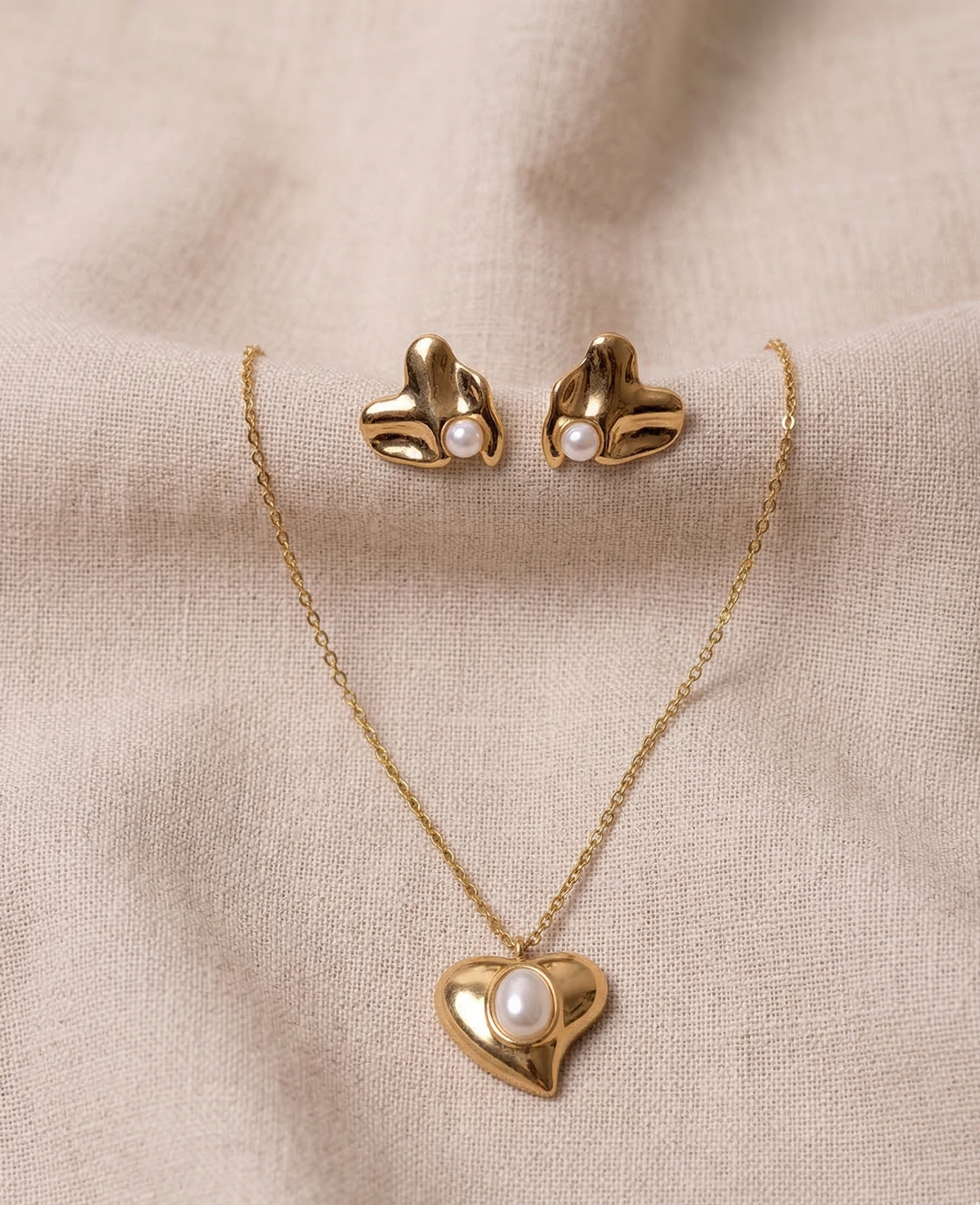 Gold heart-shaped necklace and earrings with pearl accents on a beige fabric background