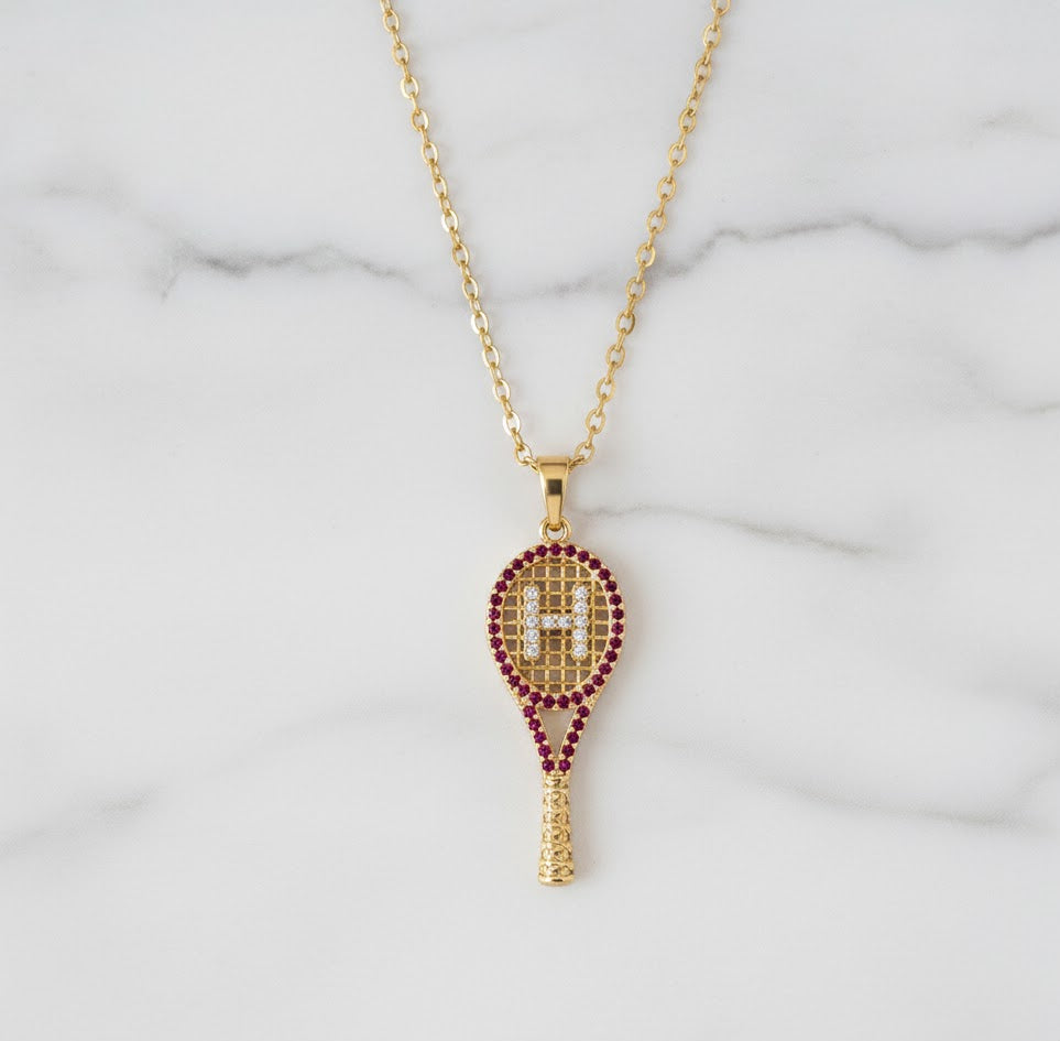 Gold tennis racket pendant necklace with red stones on a marble background