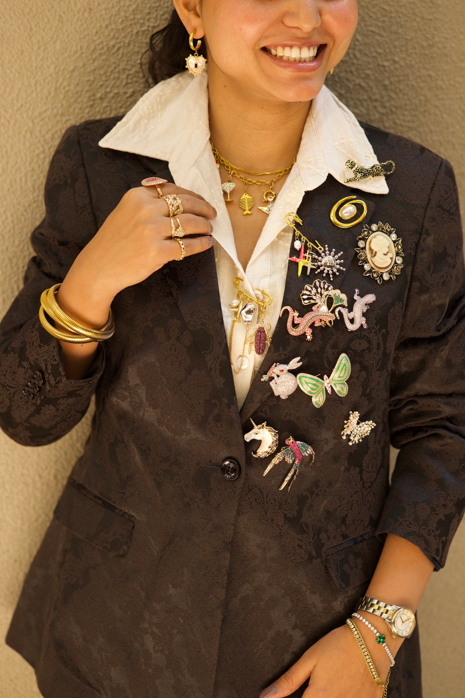 Person wearing a black blazer with various pins and jewelry against a beige wall.
