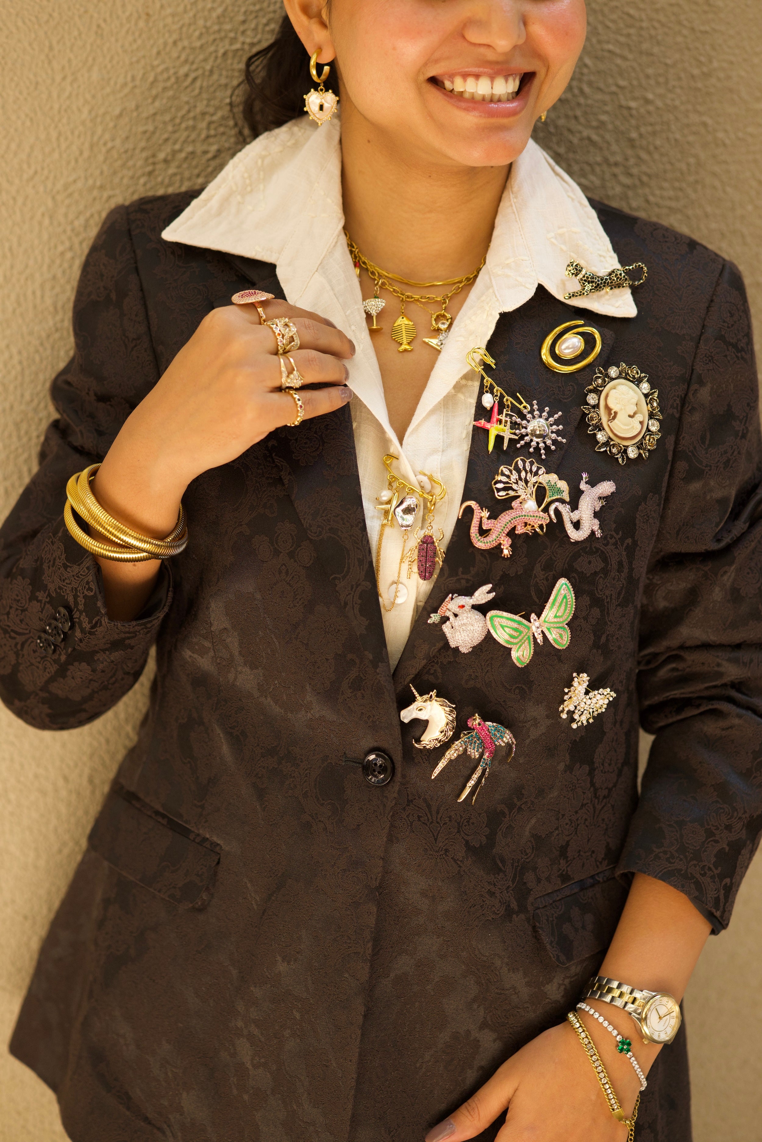 Person wearing a black blazer with various pins and jewelry against a beige wall.