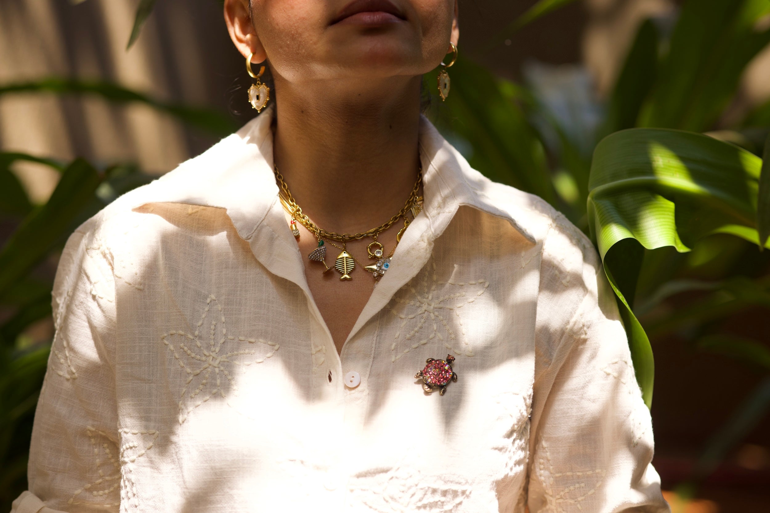 Person wearing a white shirt with jewelry and a brooch, surrounded by green foliage.