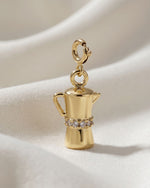 Gold charm shaped like a coffee maker with gemstones on a beige background
