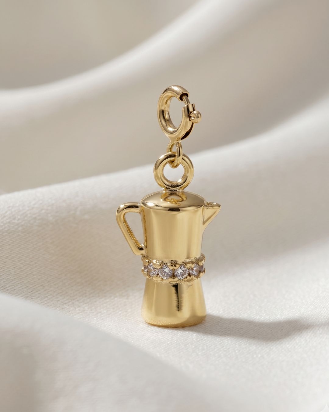 Gold charm shaped like a coffee maker with gemstones on a beige background