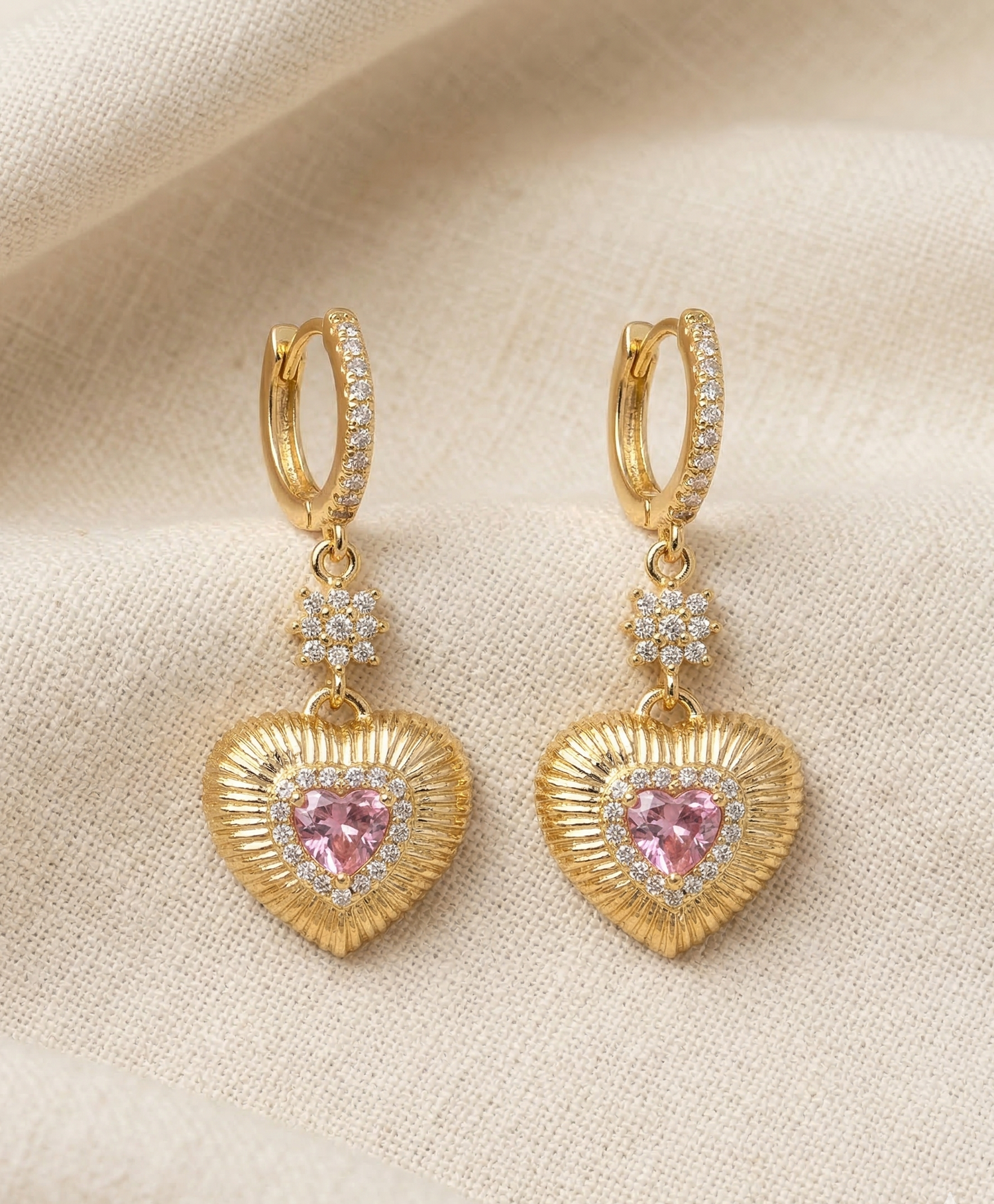 Gold heart-shaped earrings with pink gemstones on a beige fabric background