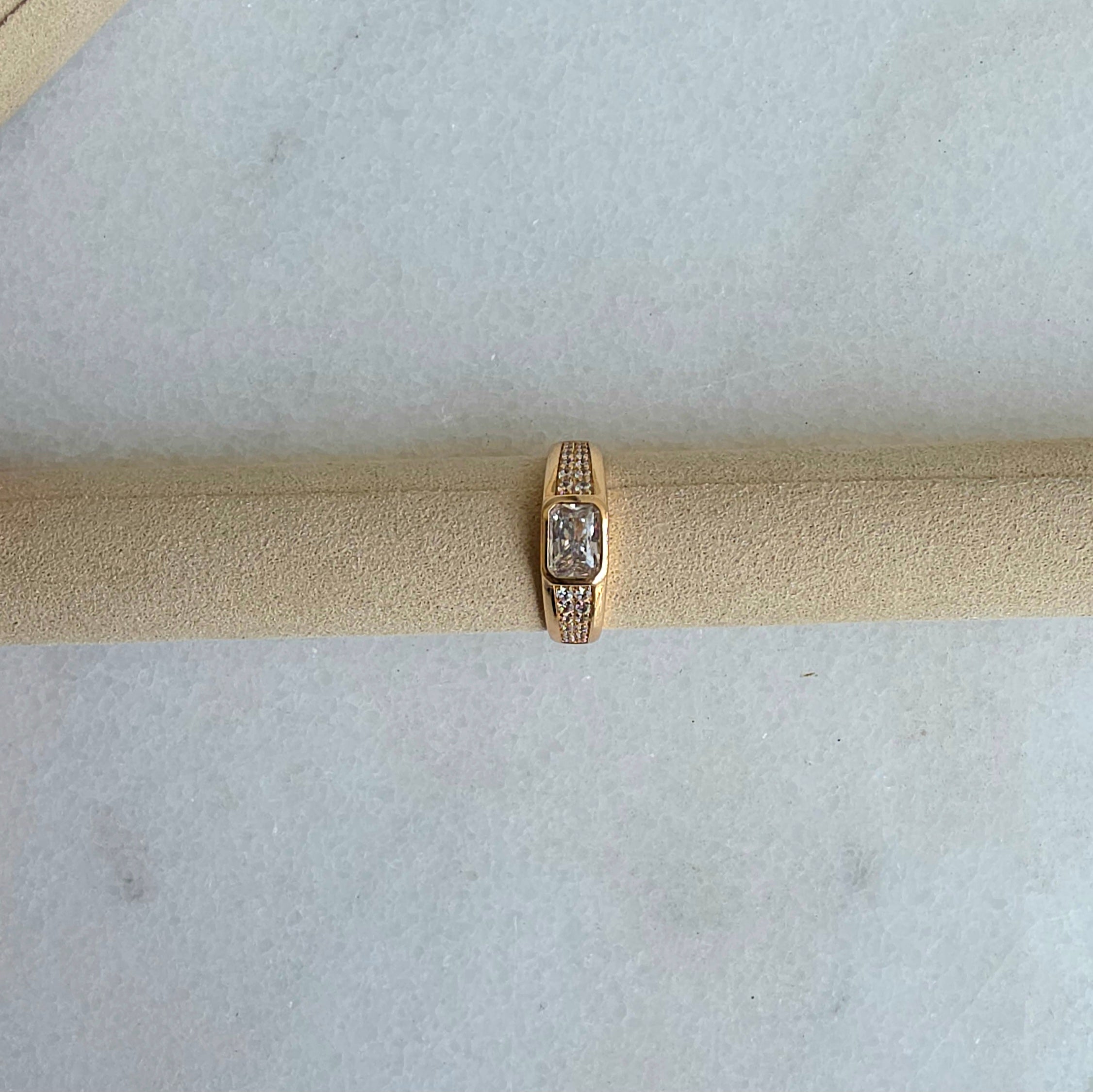 Gold ring with a diamond on a beige cushion against a light gray background