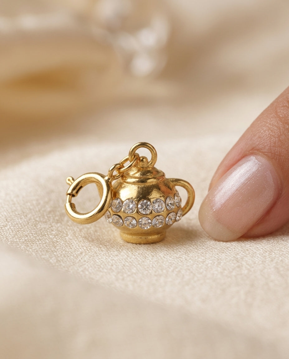 Gold teapot-shaped charm with gemstones held by a hand on a beige background