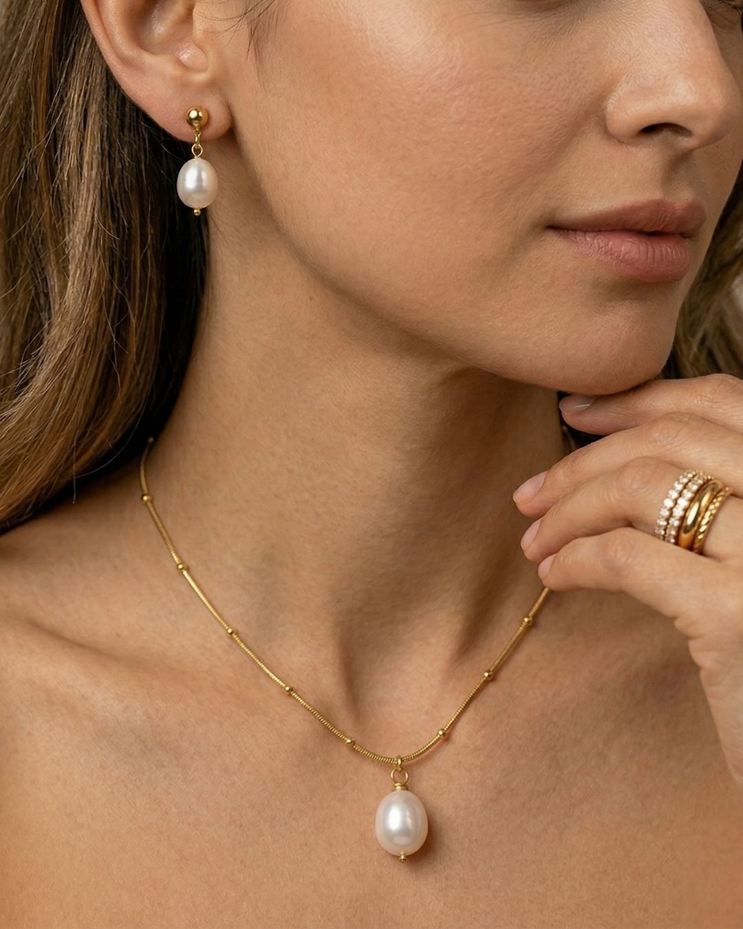 Close-up of a woman wearing a gold necklace with a pearl pendant, pearl earrings, and gold rings.