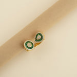 Gold earrings with green gemstones on a beige cylindrical object against a light background