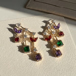 Long gold drop earrings with purple, red, emerald and amethyst halo stones stacked vertically shot on sunlight