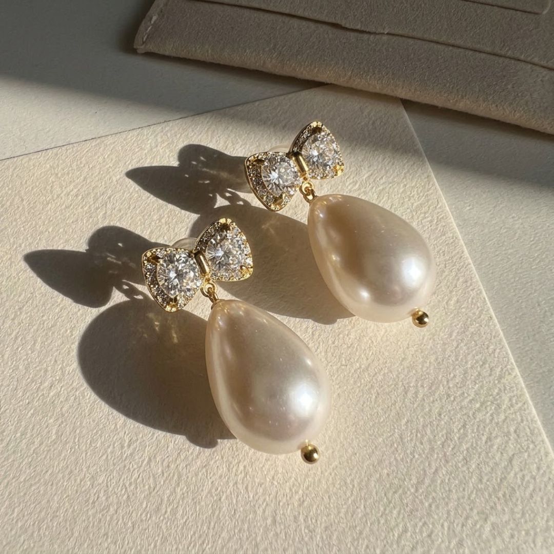 Gold CZ bow stud earrings with large teardrop baroque pearl drop held in hand against white background