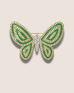 Butterfly-shaped brooch with green enamel and clear gemstones on a beige background