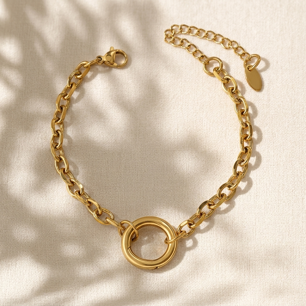 A gold chain charm bracelet with a central interlocking double-ring motif, displayed on a textured cream fabric with soft leaf shadows.