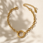 A gold chain charm bracelet with a central interlocking double-ring motif, displayed on a textured cream fabric with soft leaf shadows.