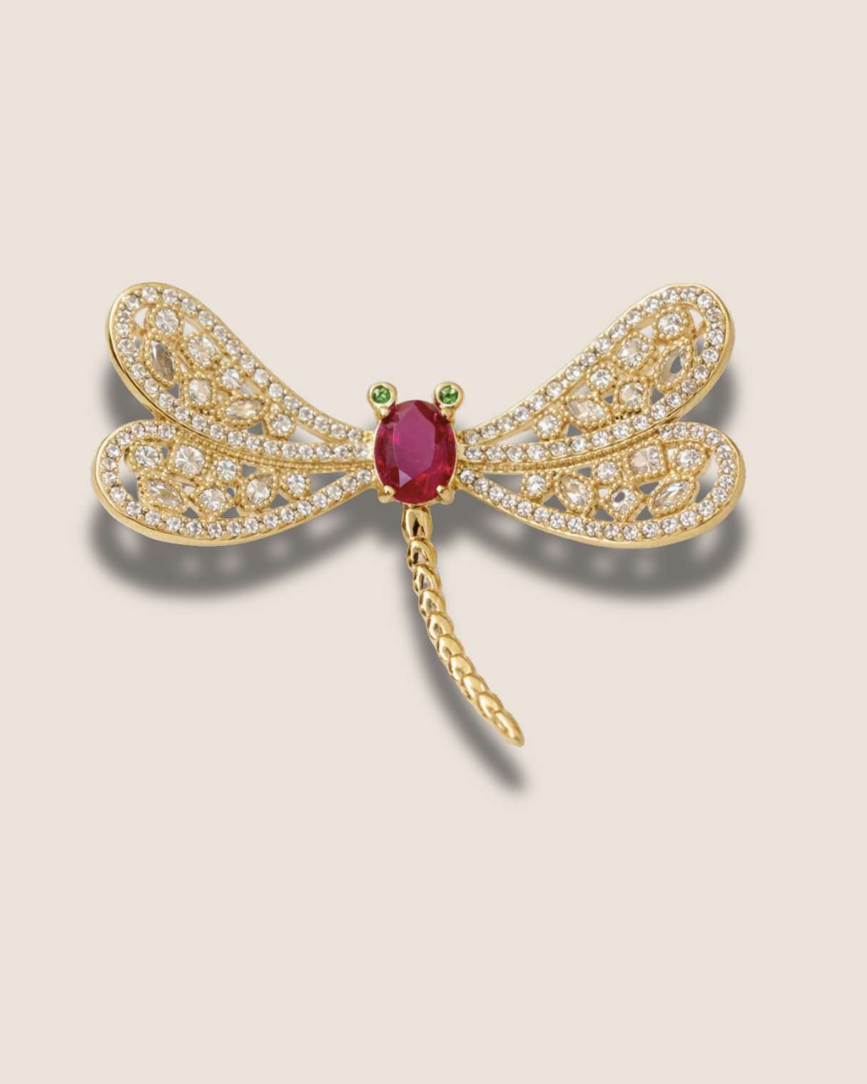 Gold dragonfly brooch with red gemstone and diamond accents on a light beige background