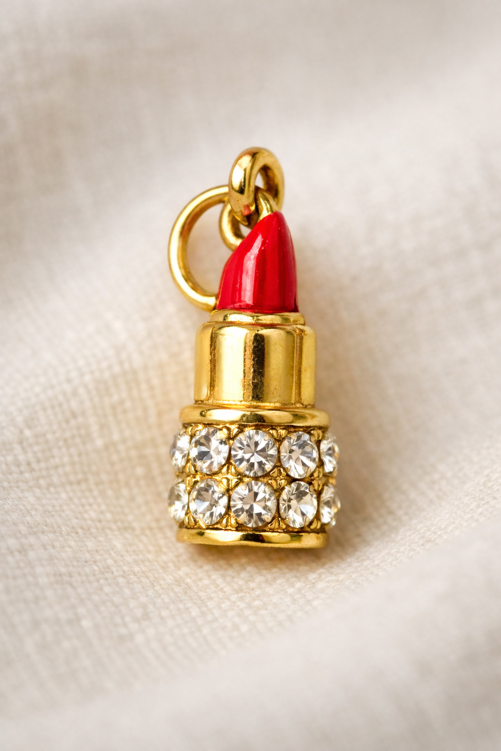 Gold lipstick charm with red top and crystal details on a beige fabric background