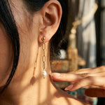 Close-up of a woman wearing a gold chain earring with a pearl drop, with a blurred background.
