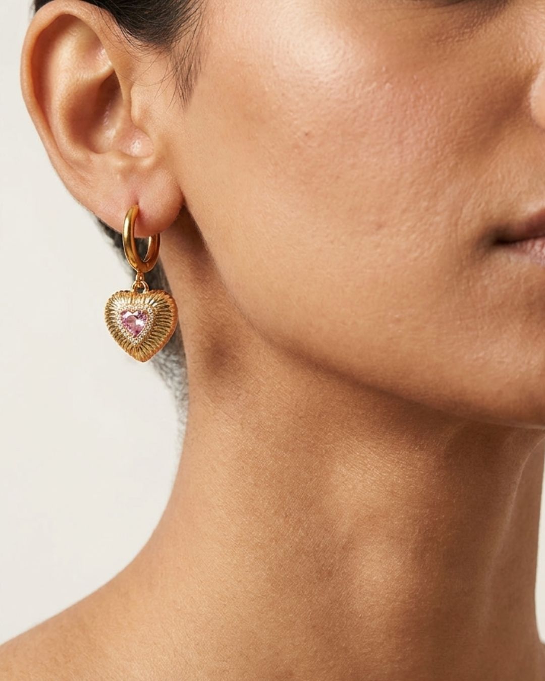 Close-up of a person wearing gold heart-shaped earrings with pink gemstones on a light background