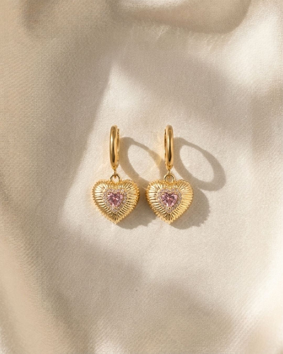 Gold heart-shaped earrings with pink gemstones on a beige background